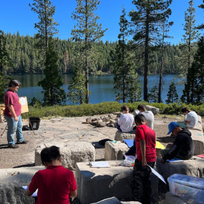 Children sitting outside near a lake, using markers to color artwork.