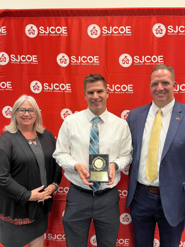 Three people in front of the SJCOE backdrop. The middle one is holding a plaque.